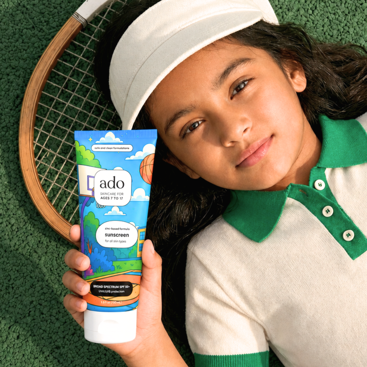Girl in a tennis visor lying on a court beside her racket, holding the ado Mineral Sunscreen SPF 50+ tube