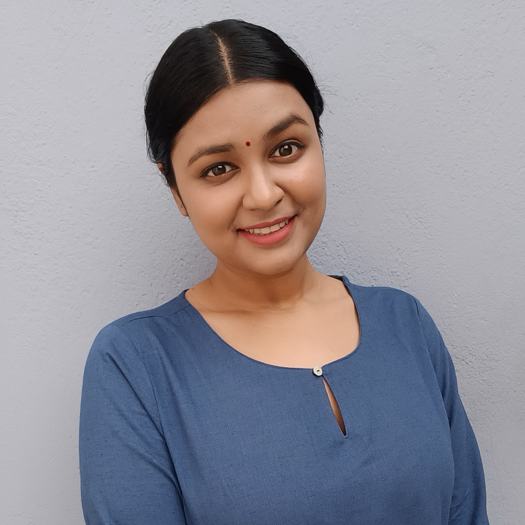 Smiling woman with her hair tied back, in a blue top against a grey wall