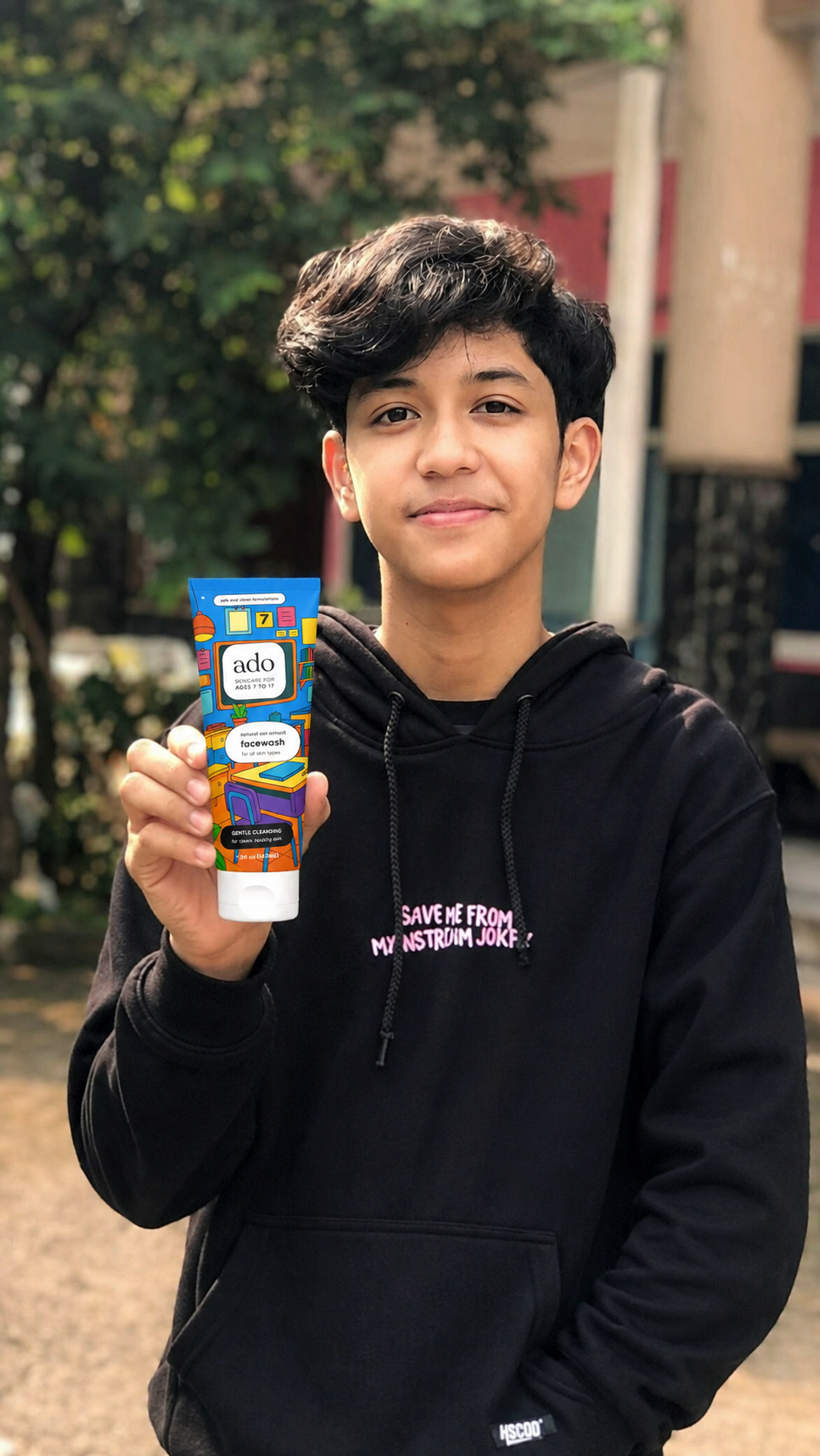 Boy in a black hoodie holding up the ado Gentle Oat Facewash tube outdoors