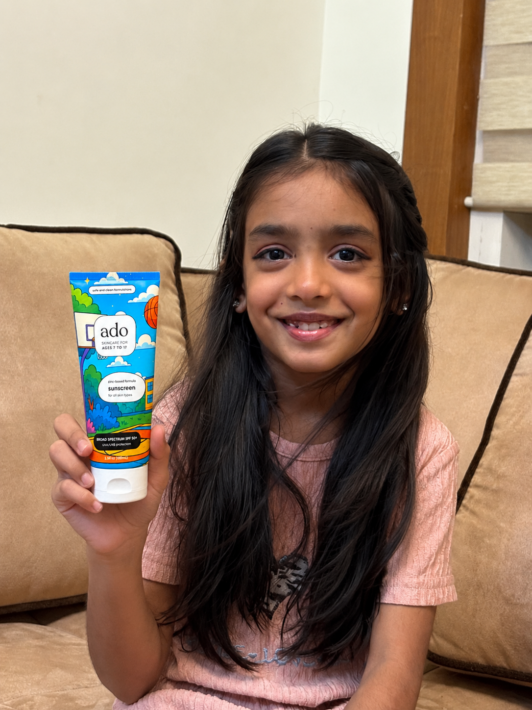 Smiling girl on a sofa holding up the ado Mineral Sunscreen SPF 50+ tube